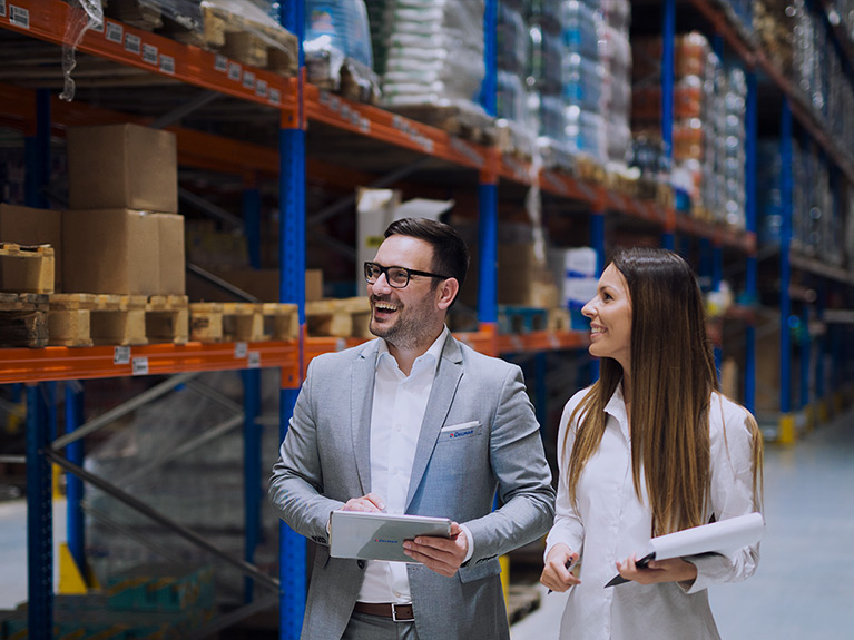 A smiling man and a smiling woman, in business attire, with clipboards in a storage warehouse, beside inventory.