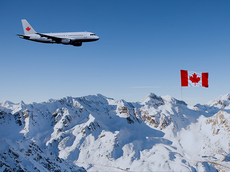 A Delmar branded airplane with a red maple leaf painted on its tailpiece in flight over a snow-covered mountain range and a Canadian flag.
