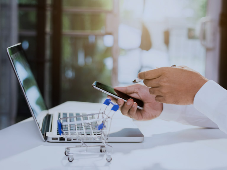 A business person holding a pen and a cellphone, in front of an open laptop computer, with a miniature shopping cart on the desk.